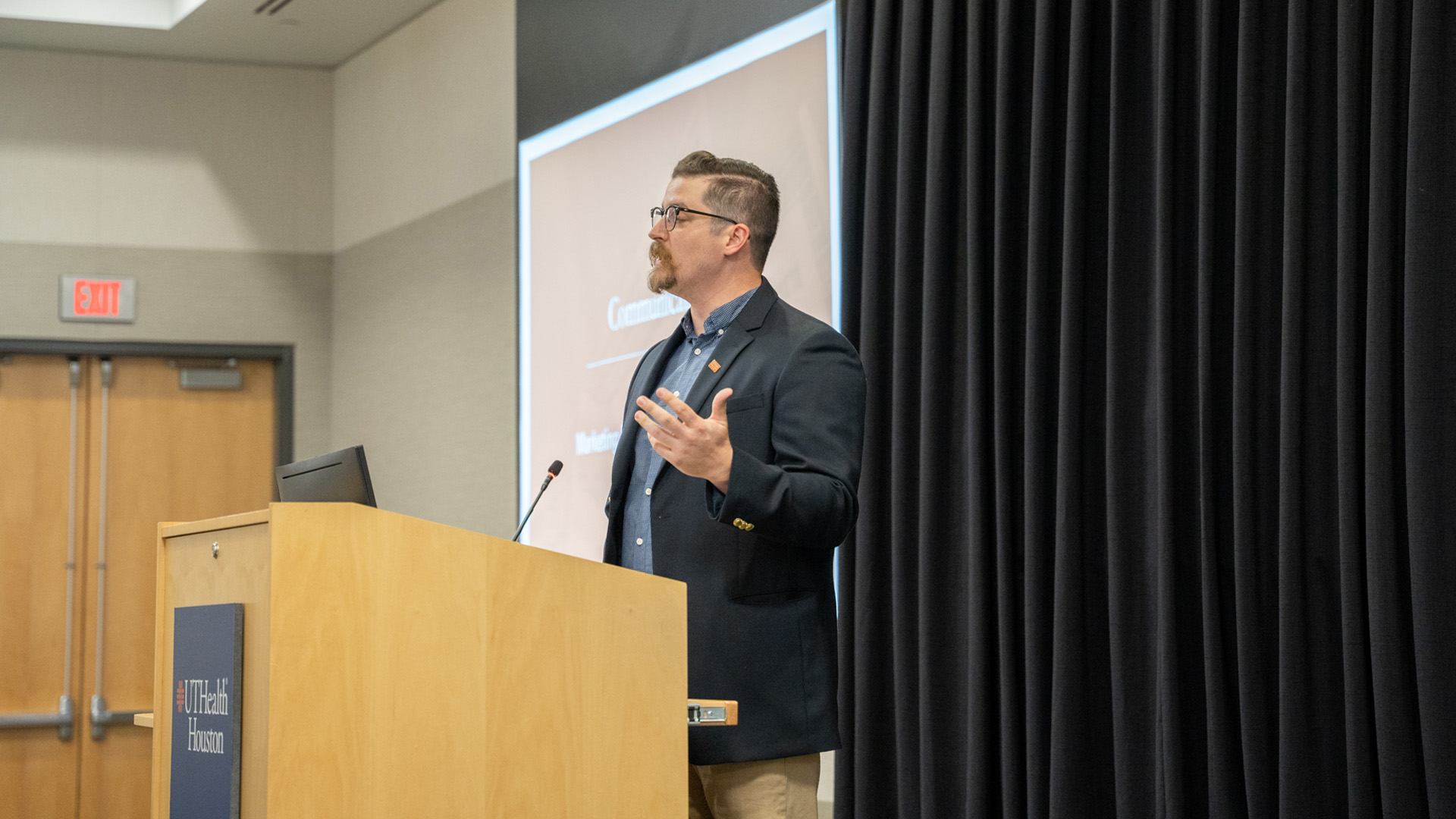 A speaker stands at a UTHealth Houston podium giving a presentation, with a large screen behind them displaying a slide in a conference setting.