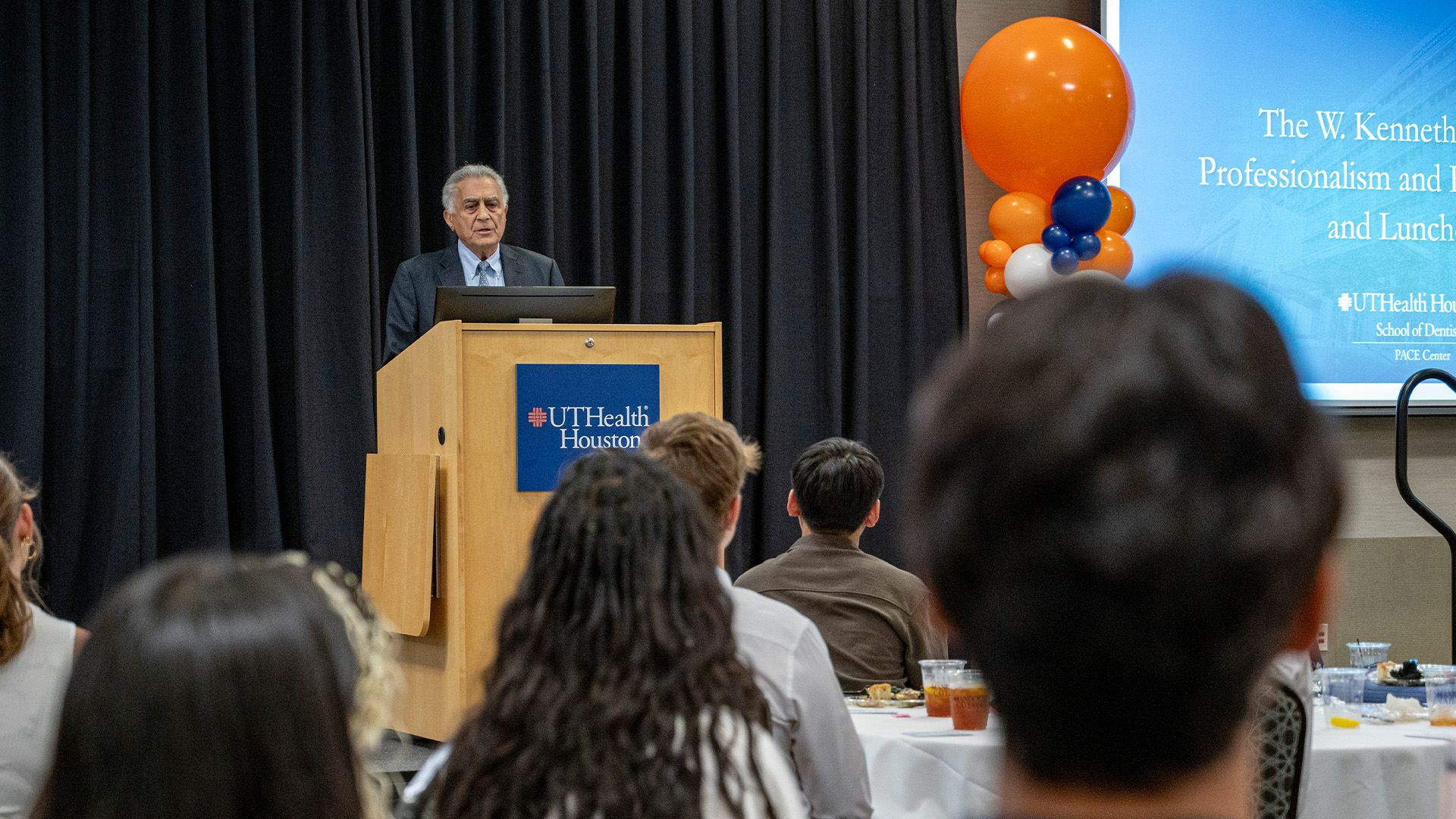 A speaker stands at a UTHealth Houston podium addressing attendees seated at round tables, with a presentation slide and orange, white, and blue balloons displayed to the side.