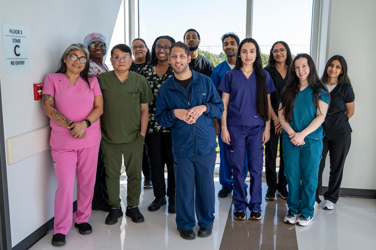 A group of men in women in scrubs stand in front of windows at the end of a hallway.