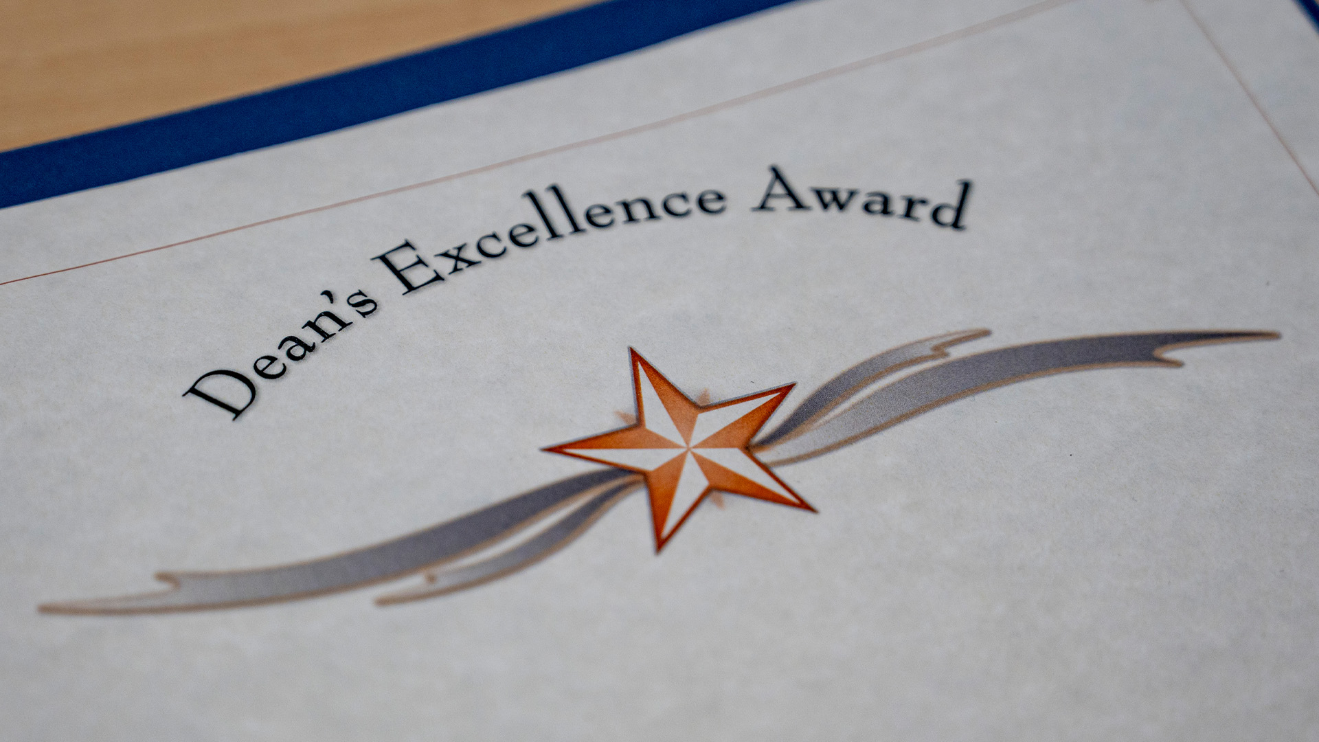 A certificate labeled Dean’s Excellence Award rests on a surface, featuring an orange and white star design with gray ribbon accents and a blue backing underneath.