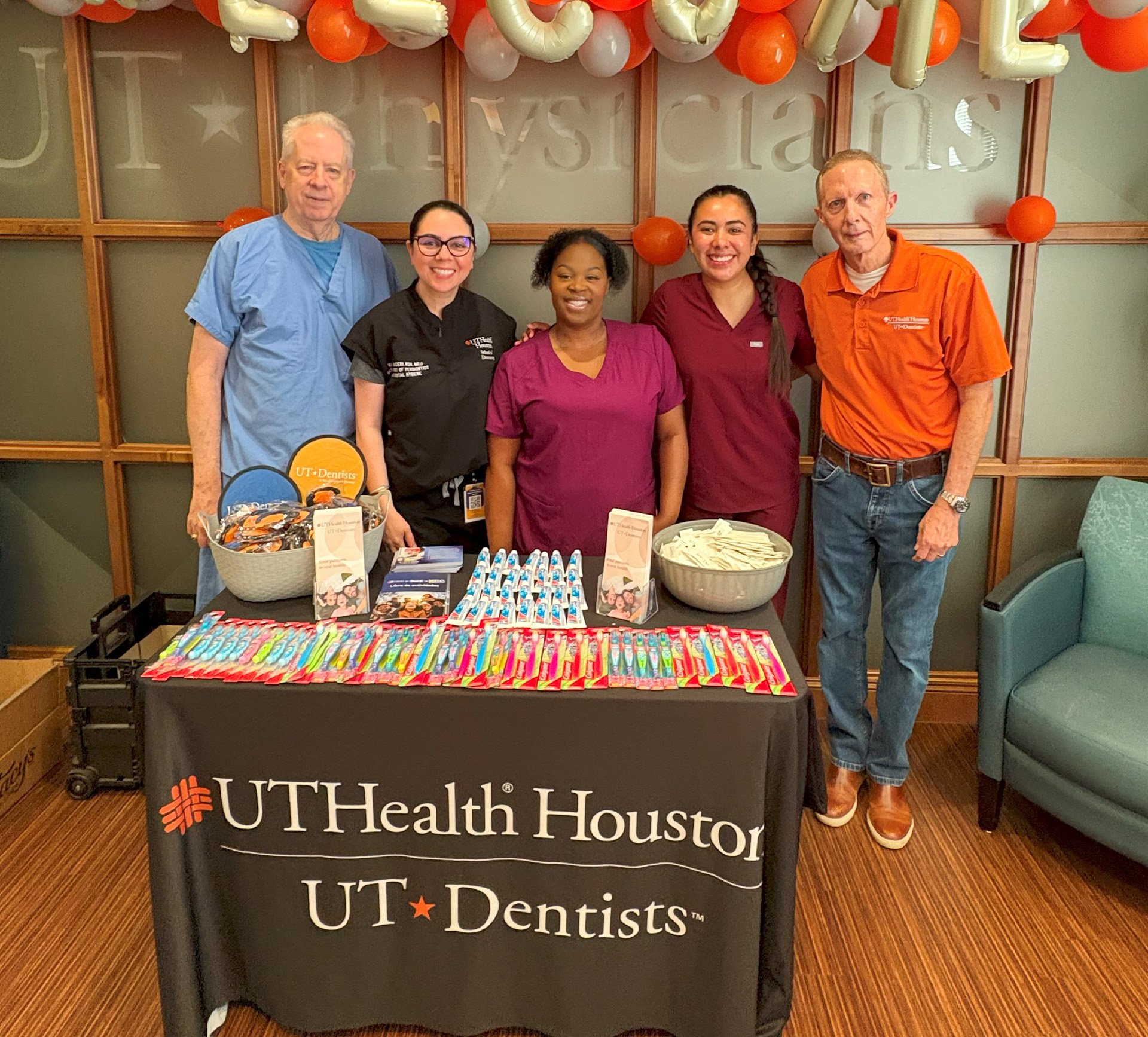 Five people stand behind a promotional table of dental hygiene products like toothbrushes, toothpaste, floss, and educational pamphlets at a community outreach event.
