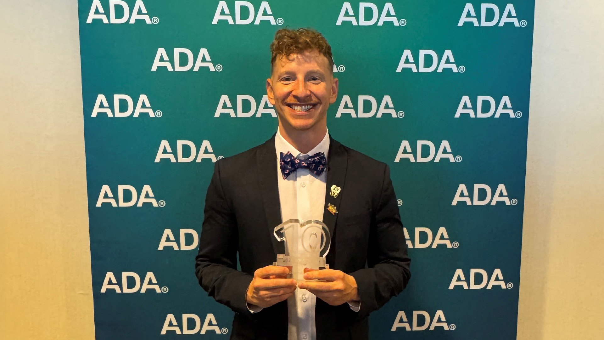 A person poses with a number 10 award in front of a backdrop displaying the ADA logo. The individual is wearing a suit and bow tie and holding the recognition award at the center of the photo.