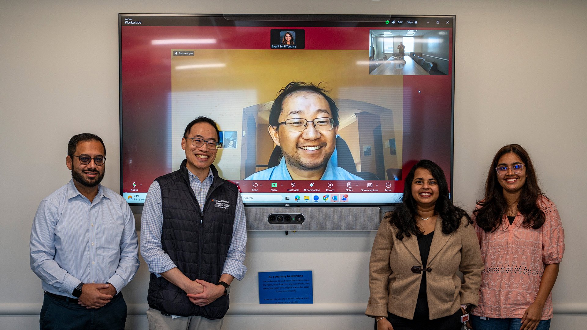 Four people stand in a conference room in front of a large wall?mounted screen displaying a video?conference call. A fifth participant appears virtually in the main video window. The in?person group smiles toward the camera as a group.