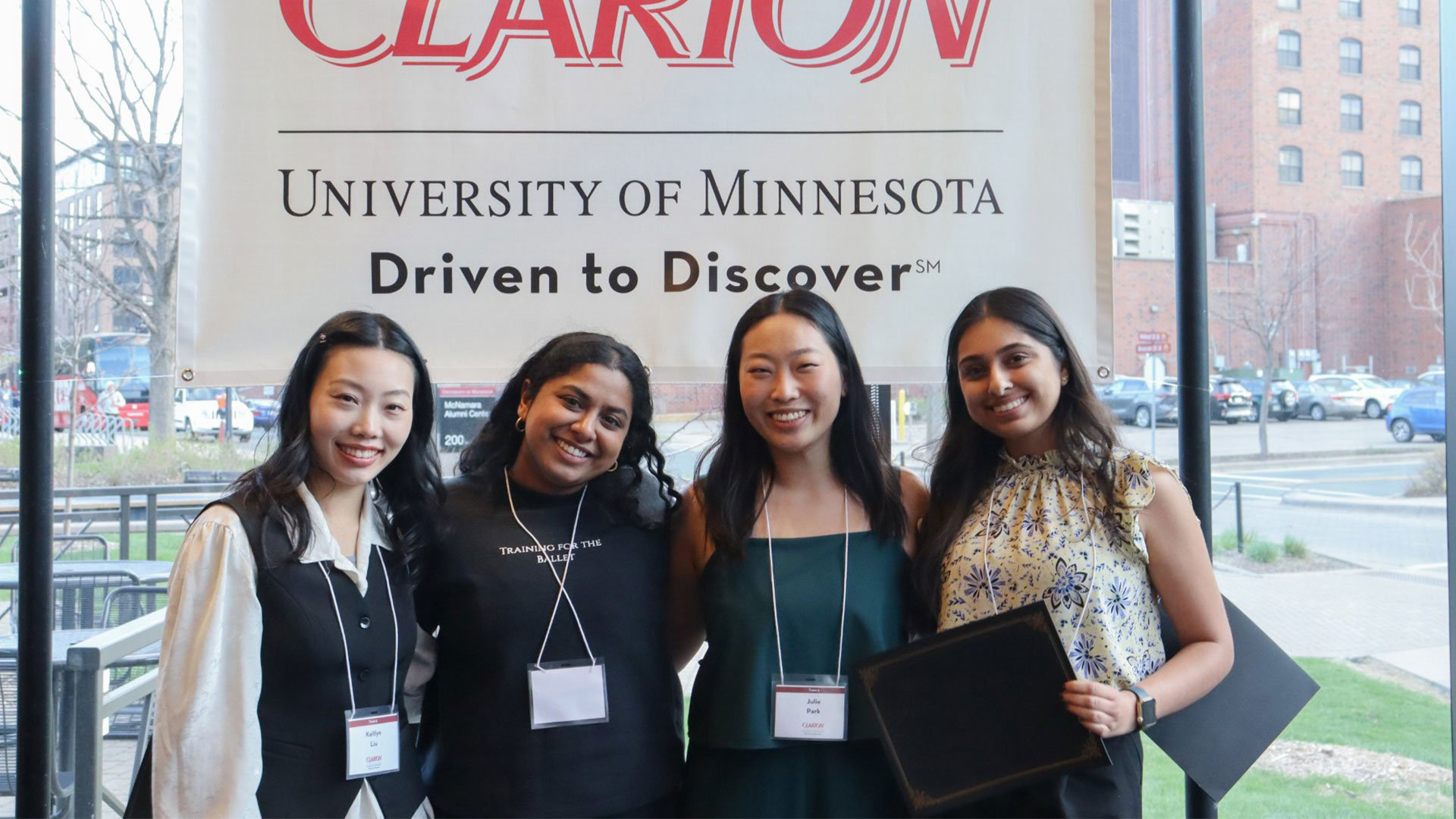 Four individuals wearing name badges stand together indoors in front of a large “CLARION” banner with city buildings visible outside behind them.