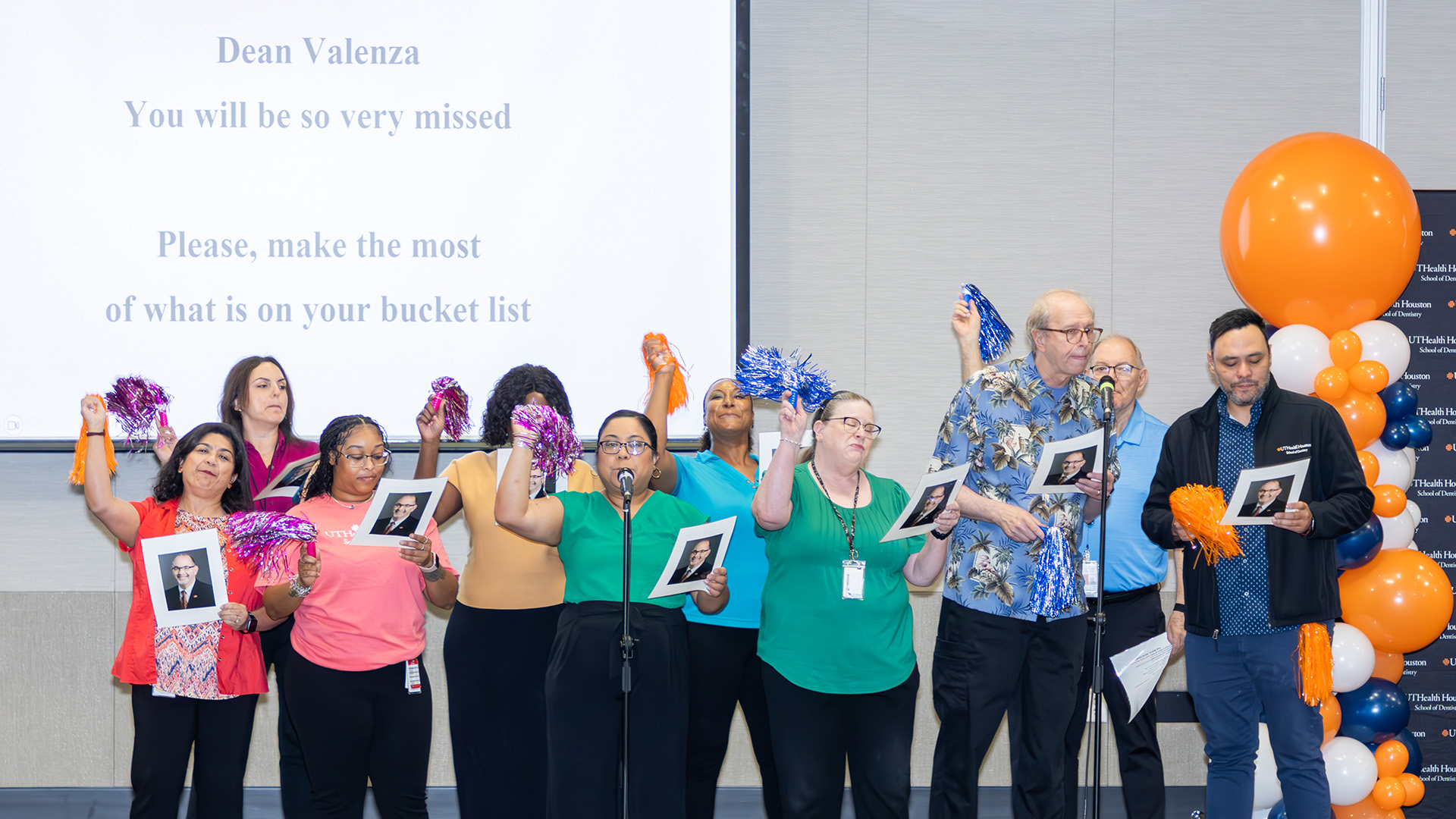 A group of people stand together at the front of a room, each holding a printed photo and colorful pom poms. Behind them, a large screen displays lyrics. A balloon arrangement frames the scene.