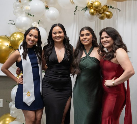 Four people in formal dresses standing in front of gold and white balloons.