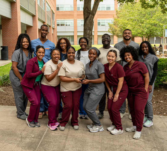 A group of people wearing maroon and gray scrubs stand together outdoors in front of a building displaying an award in the middle.