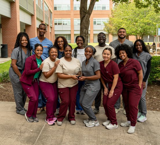 A group of people wearing maroon and gray scrubs stand together outdoors in front of a building displaying an award in the middle.