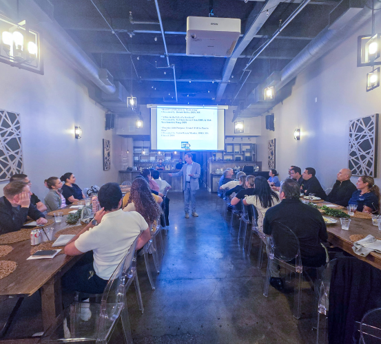 A group of people sit around two long tables in a modern, industrial-style restaurant listening to the speaker during a professional event.