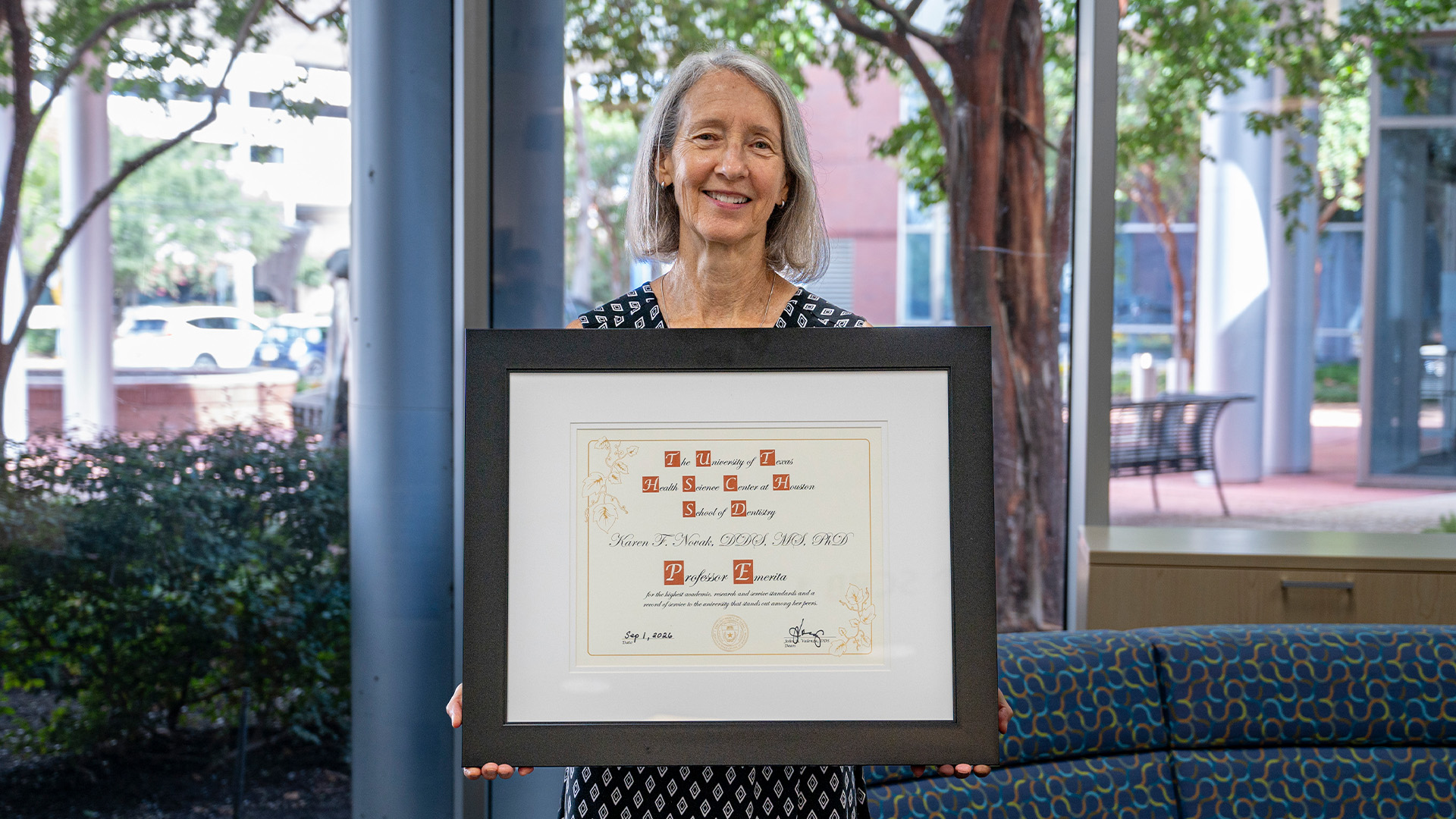 A person stands indoors holding a framed UTHealth Houston School of Dentistry certificate recognizing Karen F. Novak, DDS, MS, PhD, as professor emerita, with large windows behind them showing trees and outdoor seating.