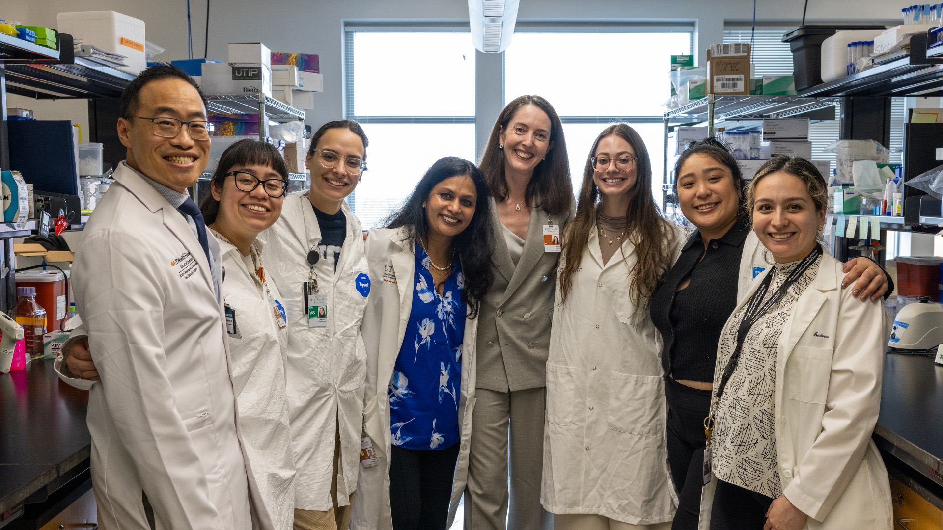 Eight individuals stand together in a research laboratory, most wearing white lab coats. They pose in front of shelves filled with lab supplies, equipment, and labeled boxes, with additional instruments and containers visible on the countertops.