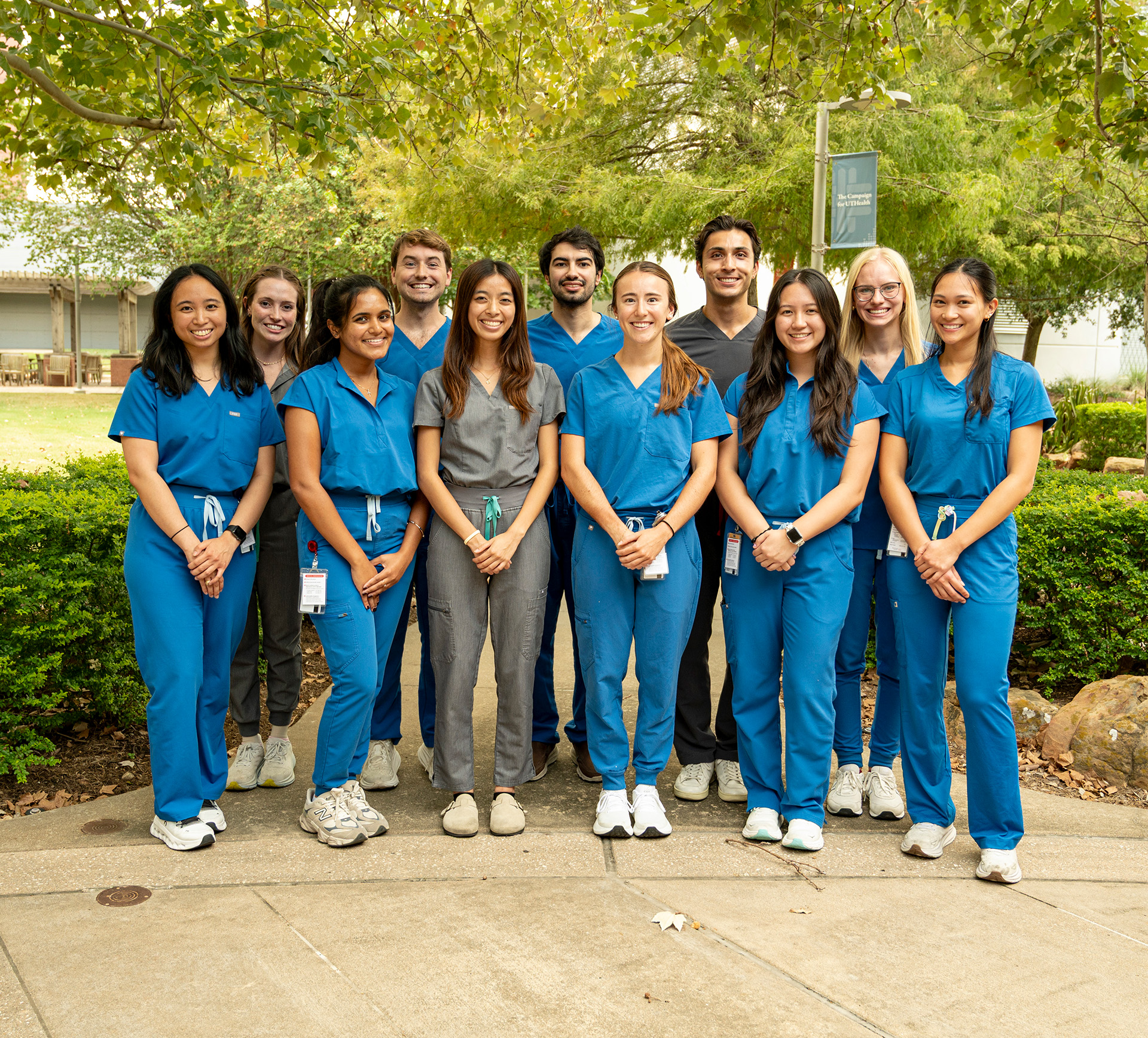 A group of eleven people, in blue scrubs and gray scrubs, pose together outdoors on a paved path surrounded by greenery.
