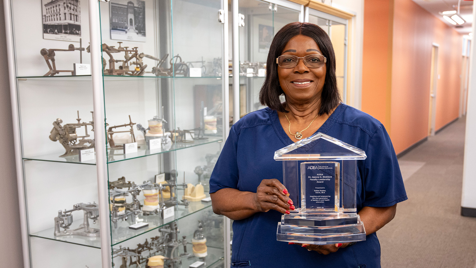A woman in blue scrubs stands in a hallway beside a glass display case filled with dental articulators. She holds a clear, house?shaped award plaque engraved with logo and award information.
