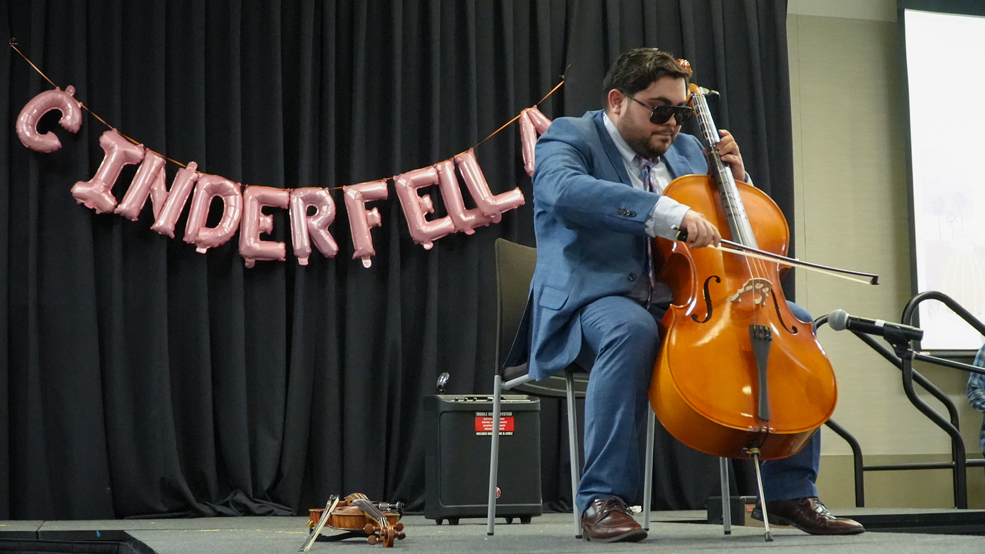 A person in a blue suit, tie, and sunglasses sits on a stage playing a cello. Pink balloon letters spelling “CINDERFELLA” hang against a black curtain behind them. A violin rests on the floor nearby.