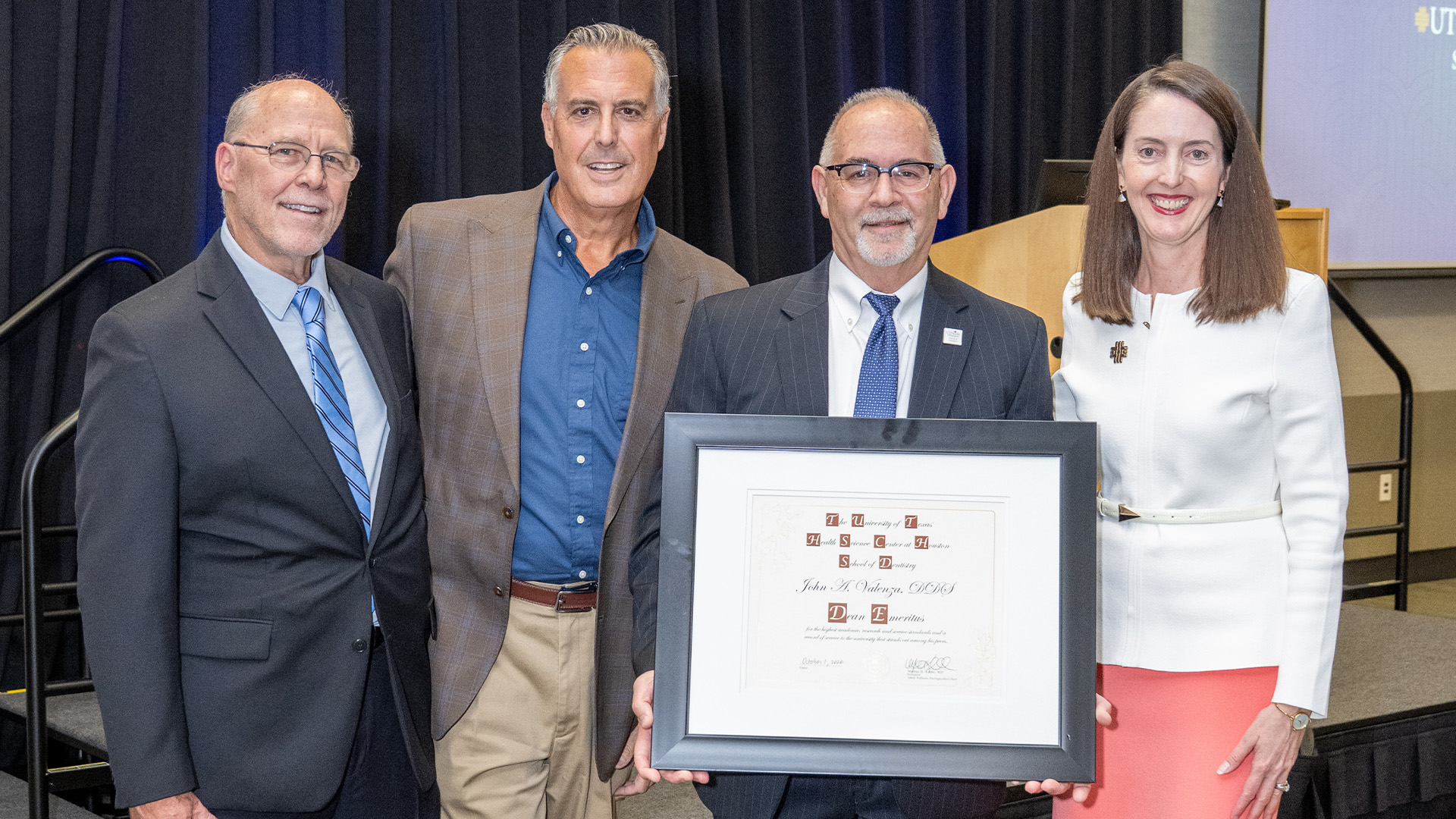 Four individuals in formal attire stand together in front of a stage as one holds a framed certificate recognizing John A. Valenza, DDS, as dean emeritus.