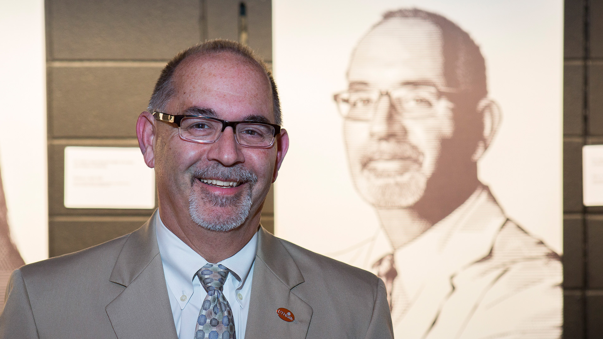 A man wearing glasses, a tan suit, and a patterned tie smiles while standing in front of a black-and-white portrait of himself on display.