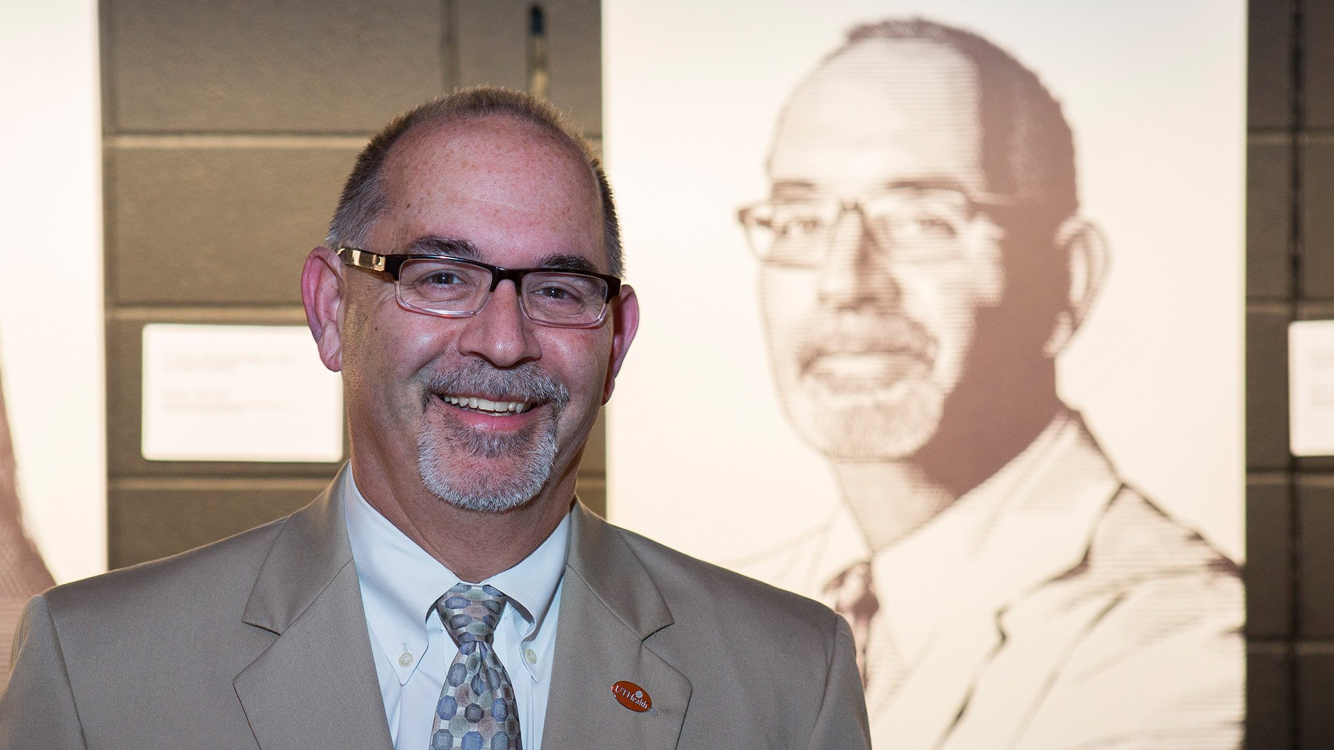 A man wearing glasses, a tan suit, and a patterned tie smiles while standing in front of a black-and-white portrait of himself on display.