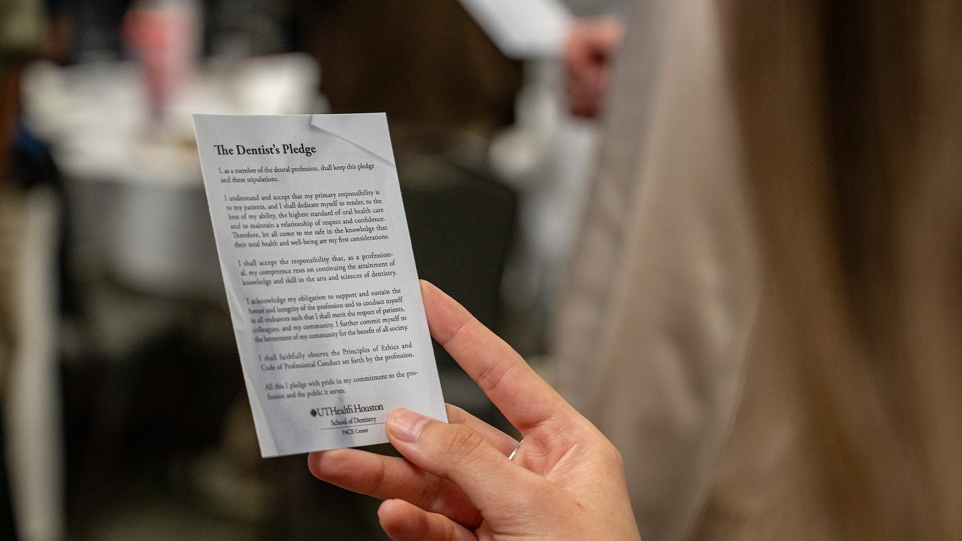 A person holds a printed card titled The Dentist’s Pledge during an indoor event, with blurred attendees and tables visible in the background.