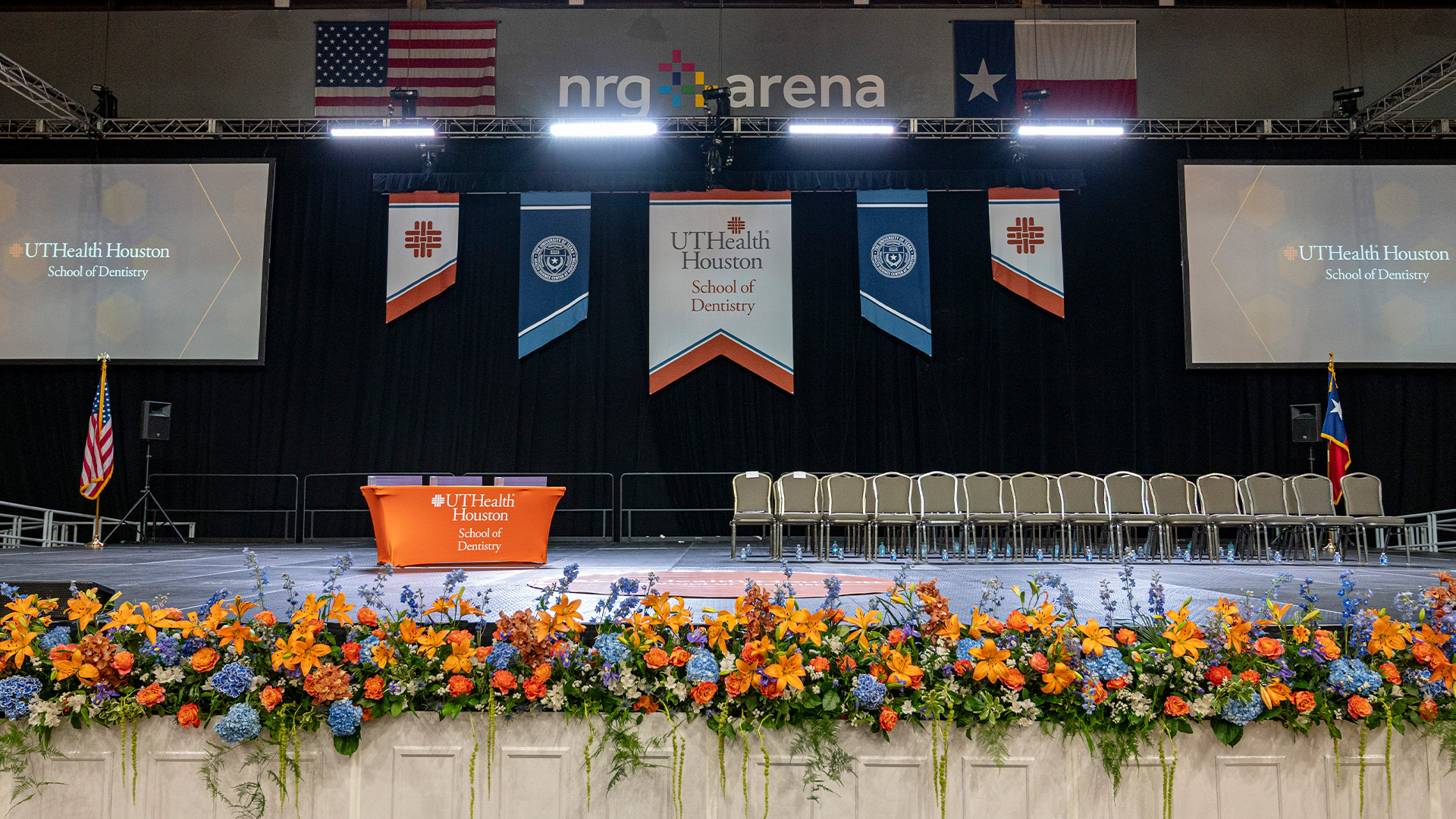 Decorated arena stage with empty chairs, floral arrangements, and hanging banners.