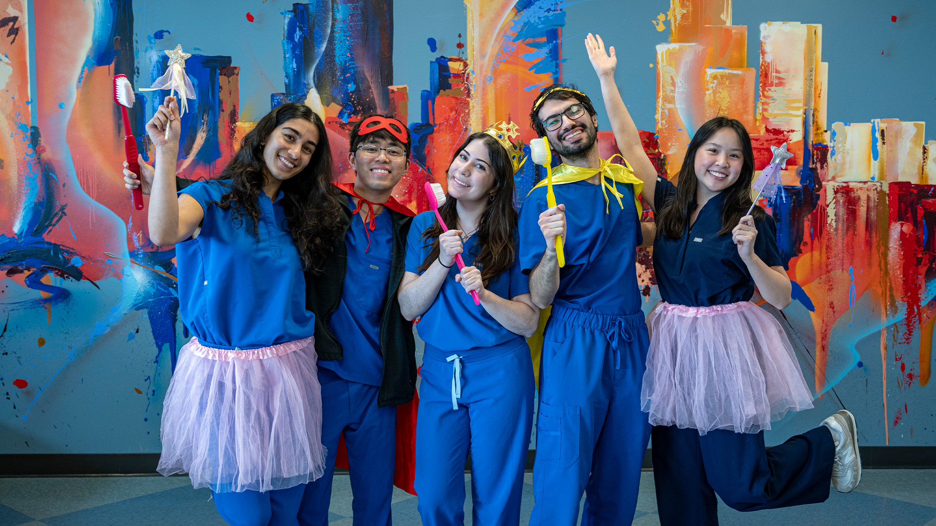 A group of five student volunteers in blue scrubs posing together in front of a colorful mural, wearing tutus and holding props.