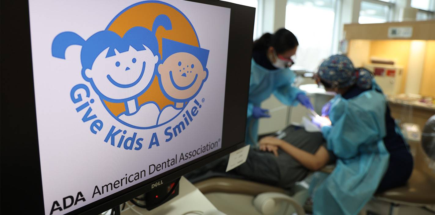 Two dental professionals attend to a patient in a dental chair. In the foreground, a computer monitor displays the Give Kids A Smile logo from the American Dental Association, featuring cartoon illustrations of a smiling boy and girl.