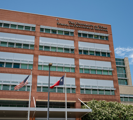 Exterior view of the UTHealth Houston School of Dentistry building, featuring a red-brick facade with rows of windows, the American and Texas flags in the foreground, and a clear blue sky overhead.