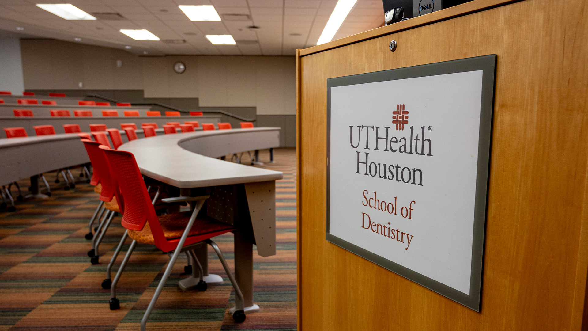 Podium displaying a UTHealth Houston School of Dentistry sign in the foreground, with rows of red seats and classroom desks in a lecture hall behind it.