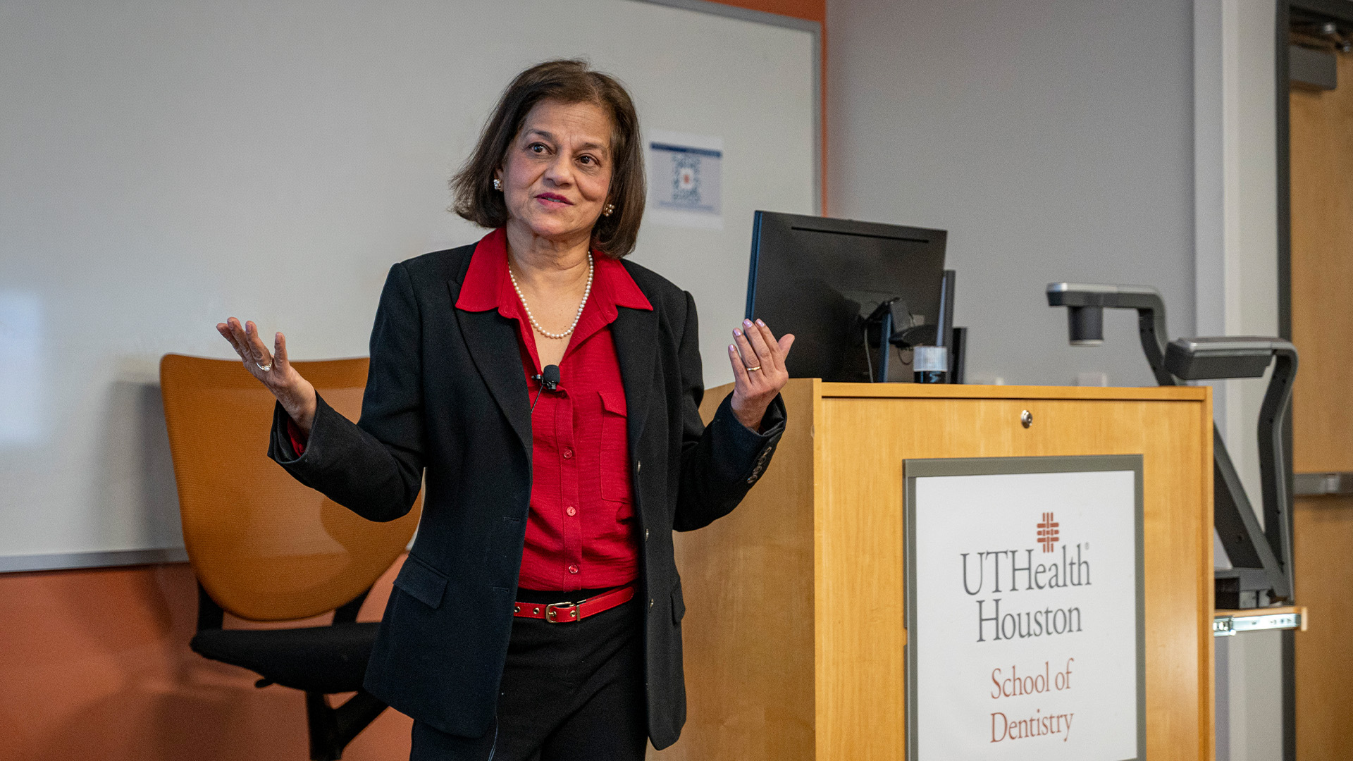 A professionally-dressed woman stands at the front of a classroom, speaking while gesturing with both hands beside a wooden podium displaying the UTHealth Houston School of Dentistry logo. A whiteboard fills the background.
