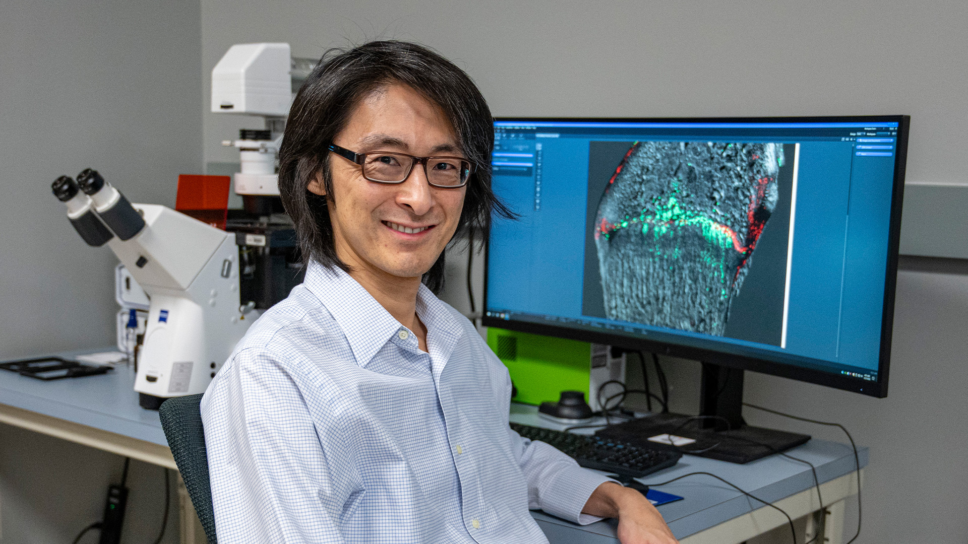 A person wearing glasses and a light blue checkered shirt sits at a desk in a laboratory. A large computer monitor beside them displays a brightly colored microscopic image with fluorescent markers. Behind the desk is a high?end microscope.