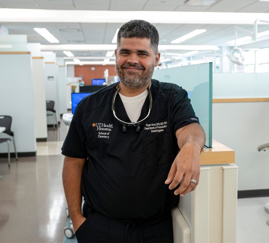 Male dentist in black scrubs leans against a clinic wall.