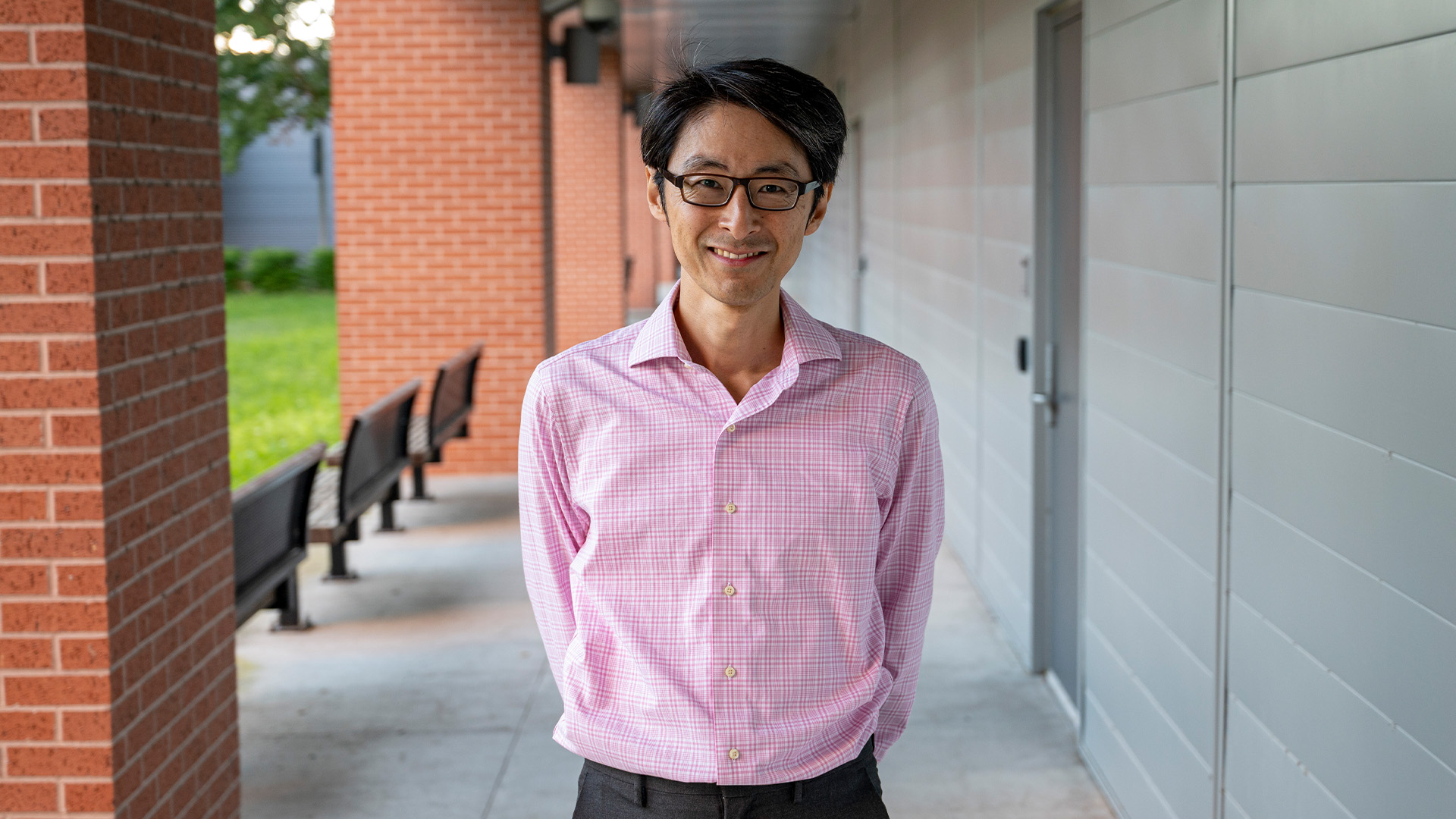 A person with short black hair and glasses stands in an outdoor corridor lined with brick columns on the left and a light gray paneled wall with a door on the right. They wear a light pink checkered button?up shirt and dark pants.