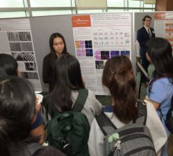 Student presents research findings from a poster to a crowd of five others.