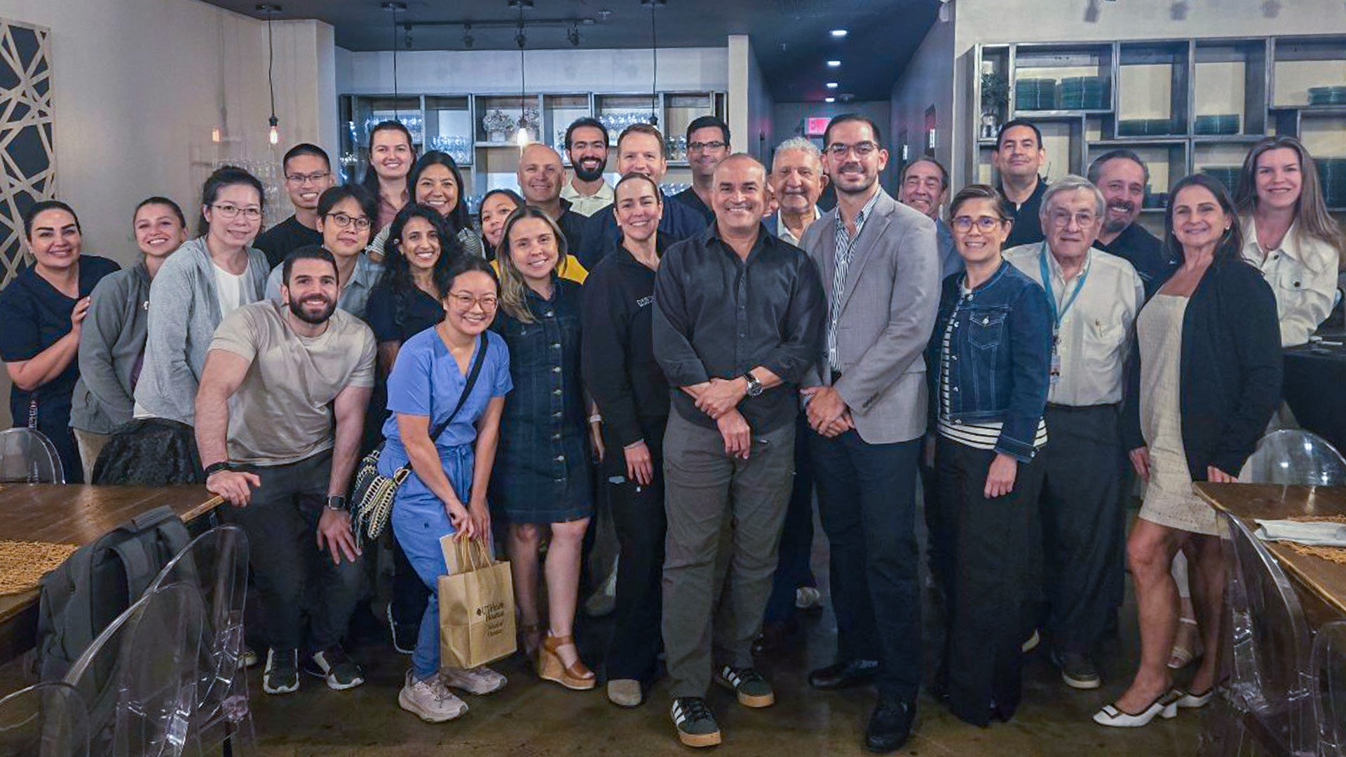 A large group of people gathers in a modern restaurant dining area, smiling at the camera at a social gathering.