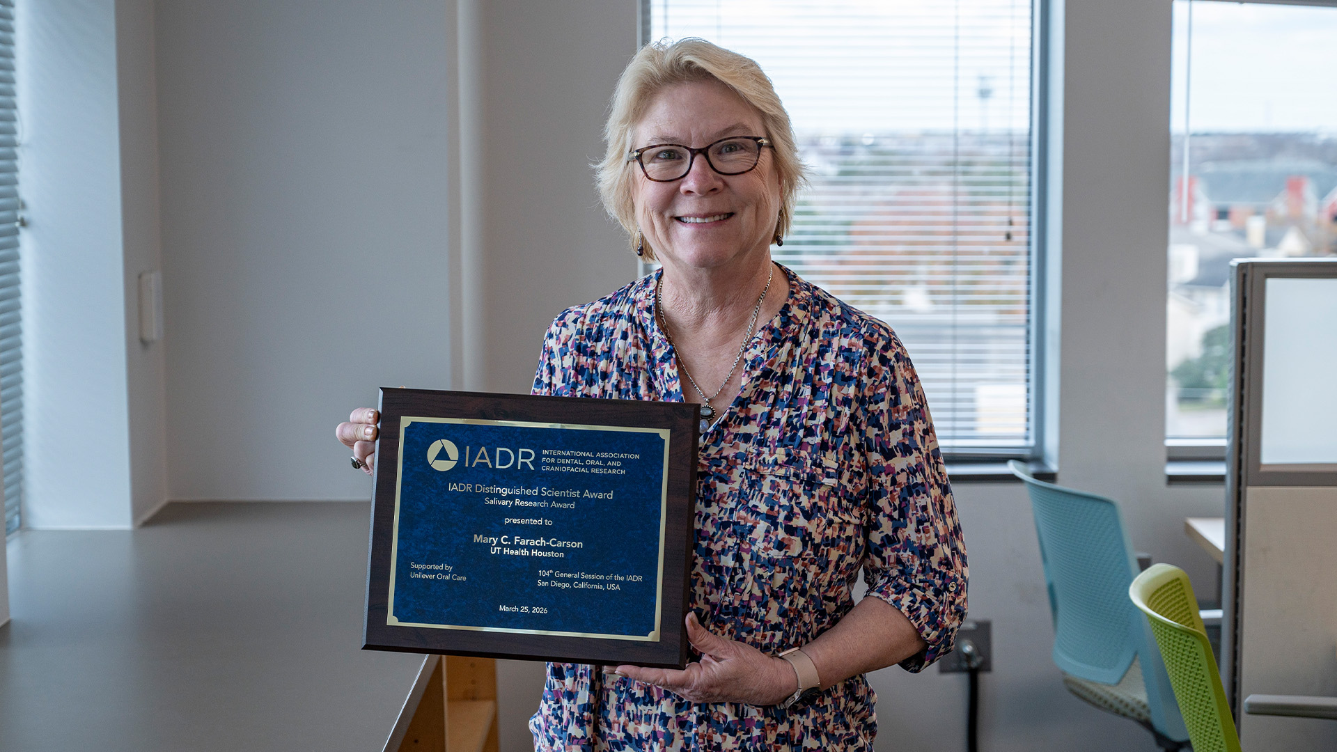 A woman wearing glasses and a patterned shirt stands in a library holding a framed award plaque. Large windows behind her show trees and nearby buildings.