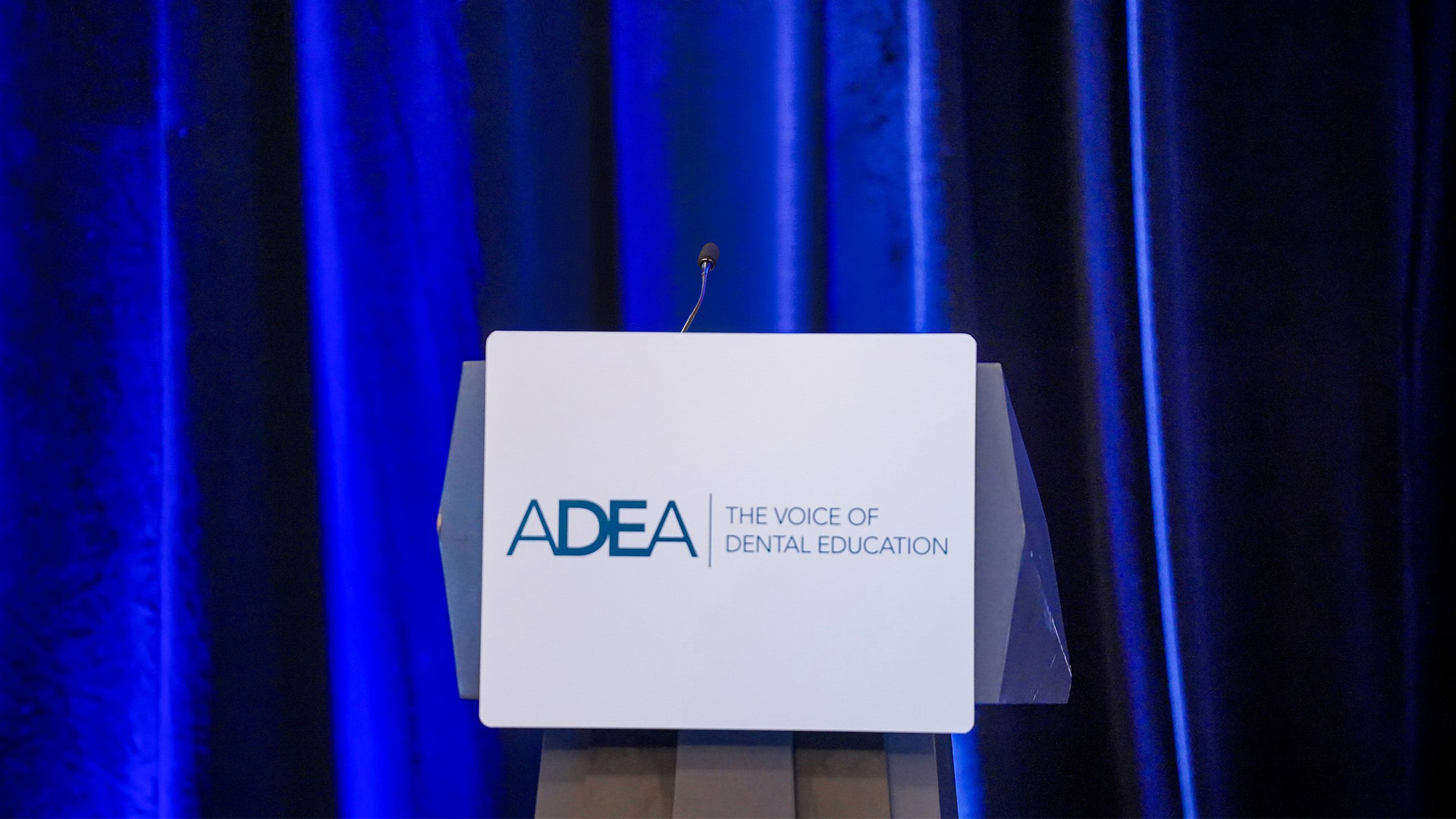 A podium with a microphone stands in front of dark blue stage curtains lit by overhead lighting. A sign on the front of the podium displays the ADEA logo and the words “The Voice of Dental Education,” indicating a formal event or conference setting.