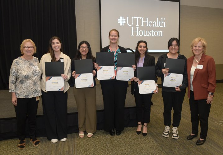 Award winners stand shoulder-to-shoulder in front of a stage for a group photo with their certificates.