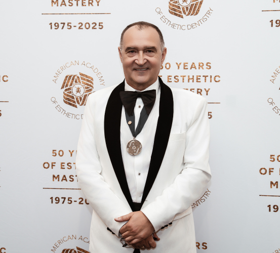 A man in a formal white tuxedo jacket with black lapels and a black bow tie wearing a medallion stands in front of a backdrop displaying the logo of the American Academy of Esthetic Dentistry.