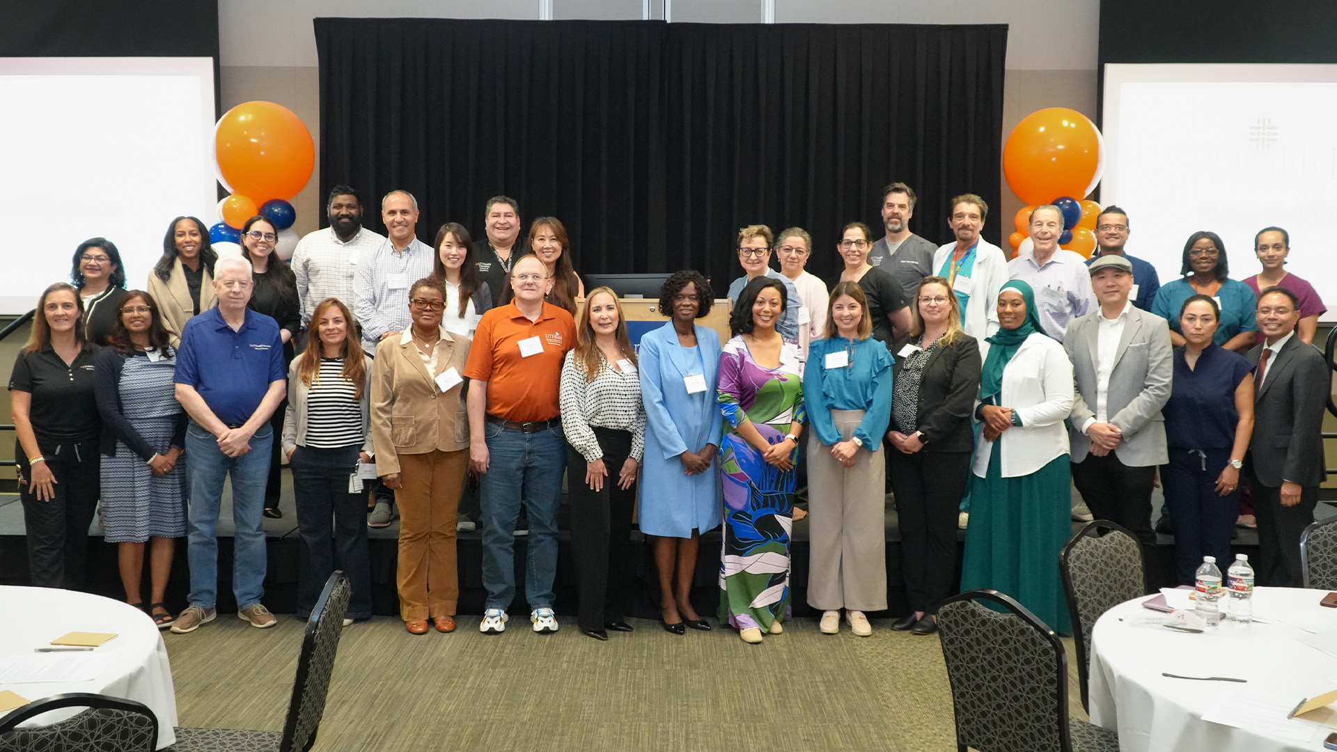 A large group of attendees pose indoors in front of a stage with a podium and black curtains, framed by orange and blue balloon arrangements, with round tables and chairs visible in the foreground.