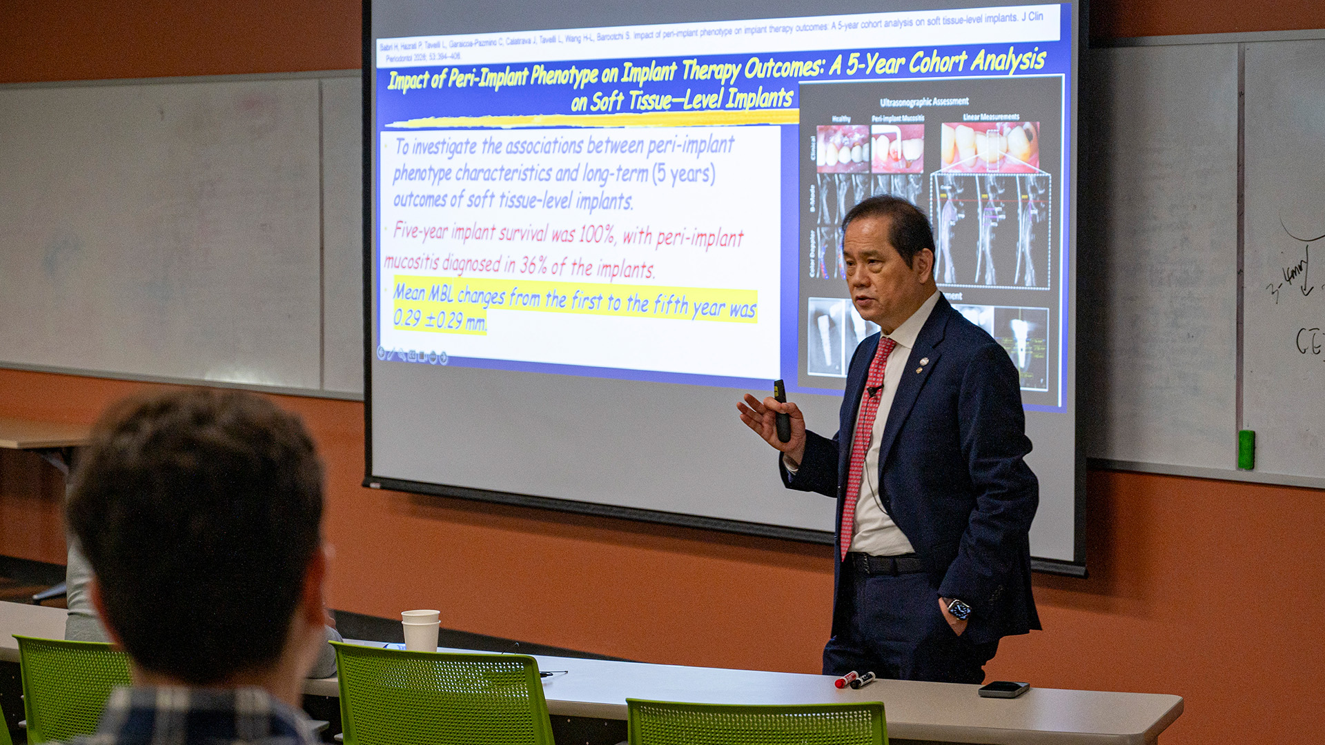 A presenter in formal attire stands at the front of a classroom, speaking beside a projected slide about peri-implant phenotype and implant therapy outcomes. A person can be seen sitting in the foreground facing his presentation.