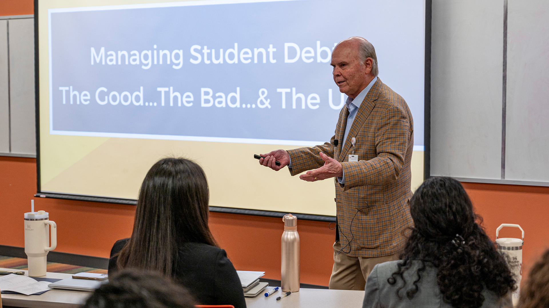 A presenter speaks beside a projected slide at the front of a classroom while two students listen from their seats.