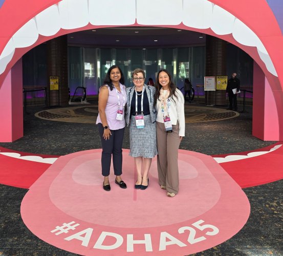 Three people standing inside a large mouth-shaped display with #ADHA25 on the floor.