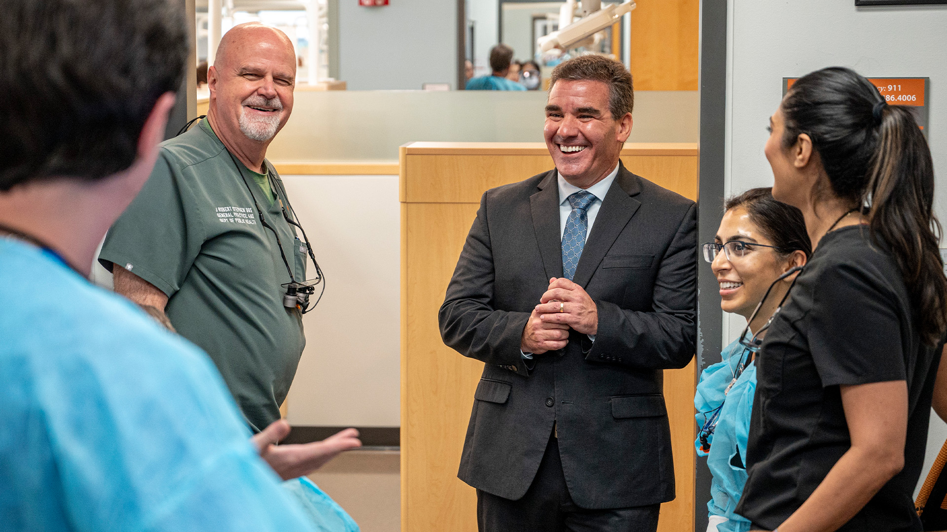 A group of five people stand in a clinical setting, with one wearing a dark suit and the others in medical scrubs, gathered near office partitions and equipment while talking together.