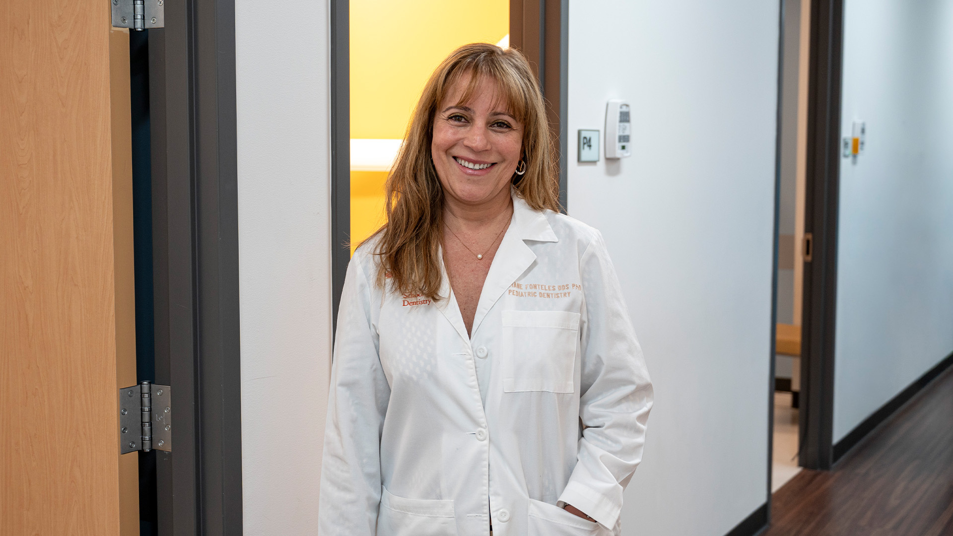 Women stands in a dental clinical hallway with her left hand in her white coat pocket.