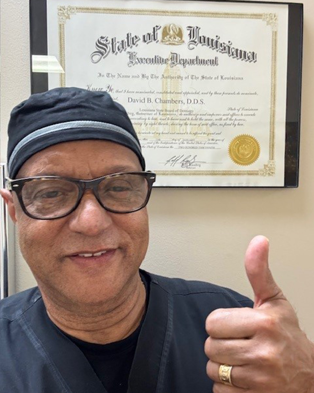 A man wearing glasses and medical scrubs gives a thumbs-up while standing in front of a framed Louisiana state certificate on the wall.