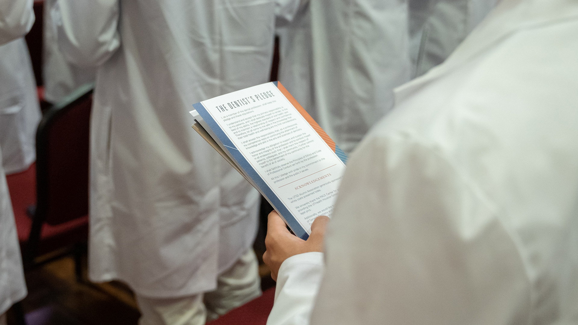 A person in a white coat reads The Dentist’s Pledge from the back of a program during a formal ceremony.