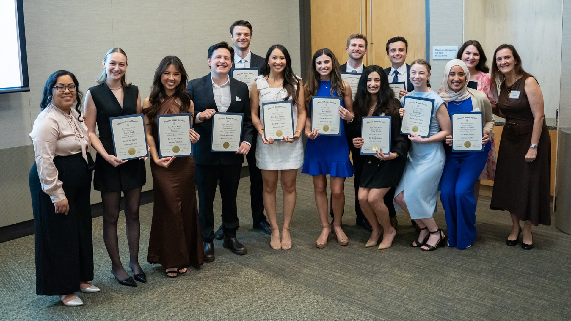A group of people stand indoors holding certificates during a recognition event associated with UTHealth Houston School of Dentistry. They are dressed in formal attire and pose in front of wooden double doors and a projection screen.