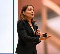 A woman in a black blazer speaks into a handheld microphone while holding a presentation remote standing in front of a large screen video screen.