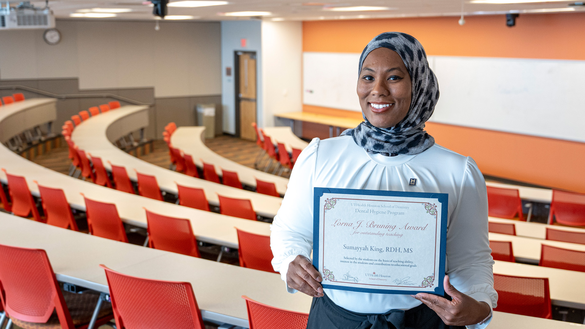 Person holding a framed certificate while standing in a tiered classroom with rows of red seats and curved desks.