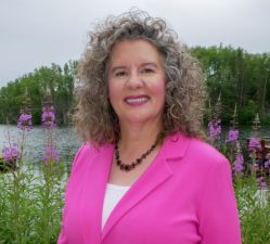 Women in pink blazer stands near flowers in front of a body of water and trees.
