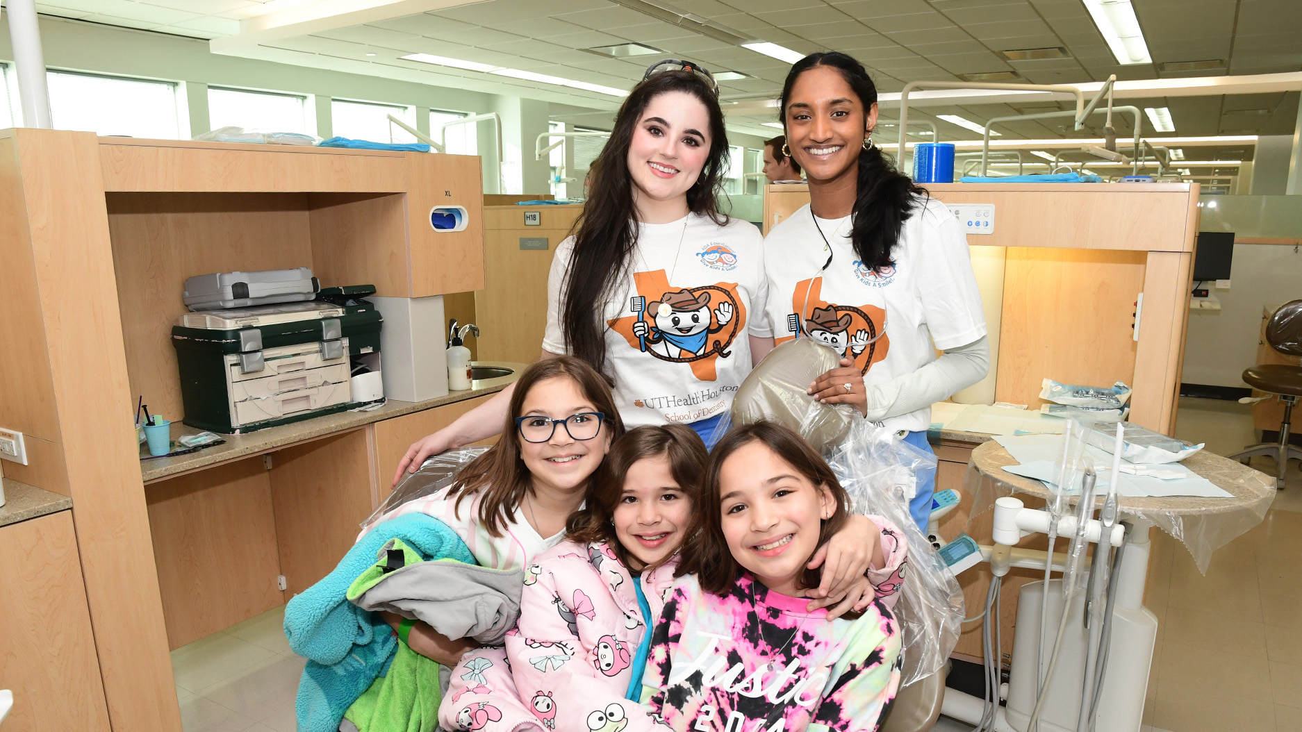 Two dental students stand behind a trio of kid sisters inside a dental operatory in a clinical setting.